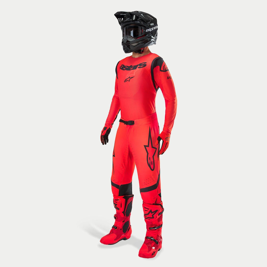 Motocyklista motocrossowy w kompletnym stroju wyścigowym, koszulka i spodnie Supertech Ember LE w kolorystyce Red Fluo i czarnej, czarny kask z goglami, pasujące czerwono-czarne buty i rękawice, profesjonalny zestaw do wyścigów off-road